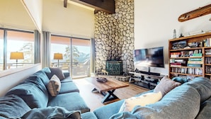 A cozy living room with a stone wall, a large TV, a bookshelf, and a sofa. - Deluxe Home, Great Views! Private Gas Grill & Washer/Dryer, Near Lakes Basin (Mammoth Lakes)