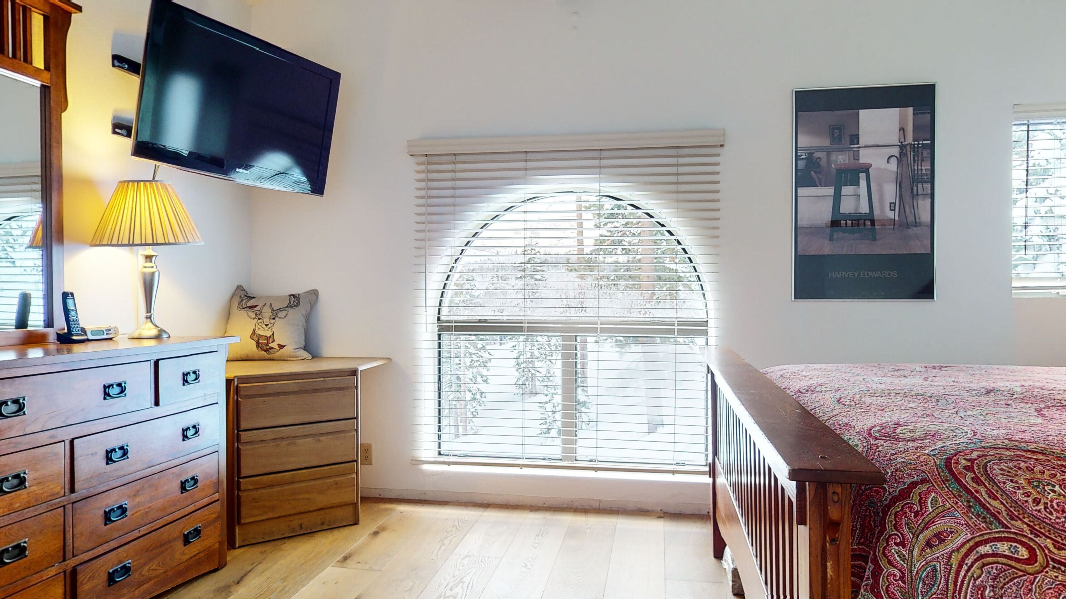 A bedroom with a wooden dresser, a television mounted on the wall, a window with blinds, and a poster on the wall.