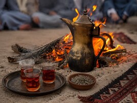 Una disposición tradicional para el té, con un hervidor metálico, tres vasos de té y un cuenco de hierbas sobre una bandeja, con un fuego como fondo.