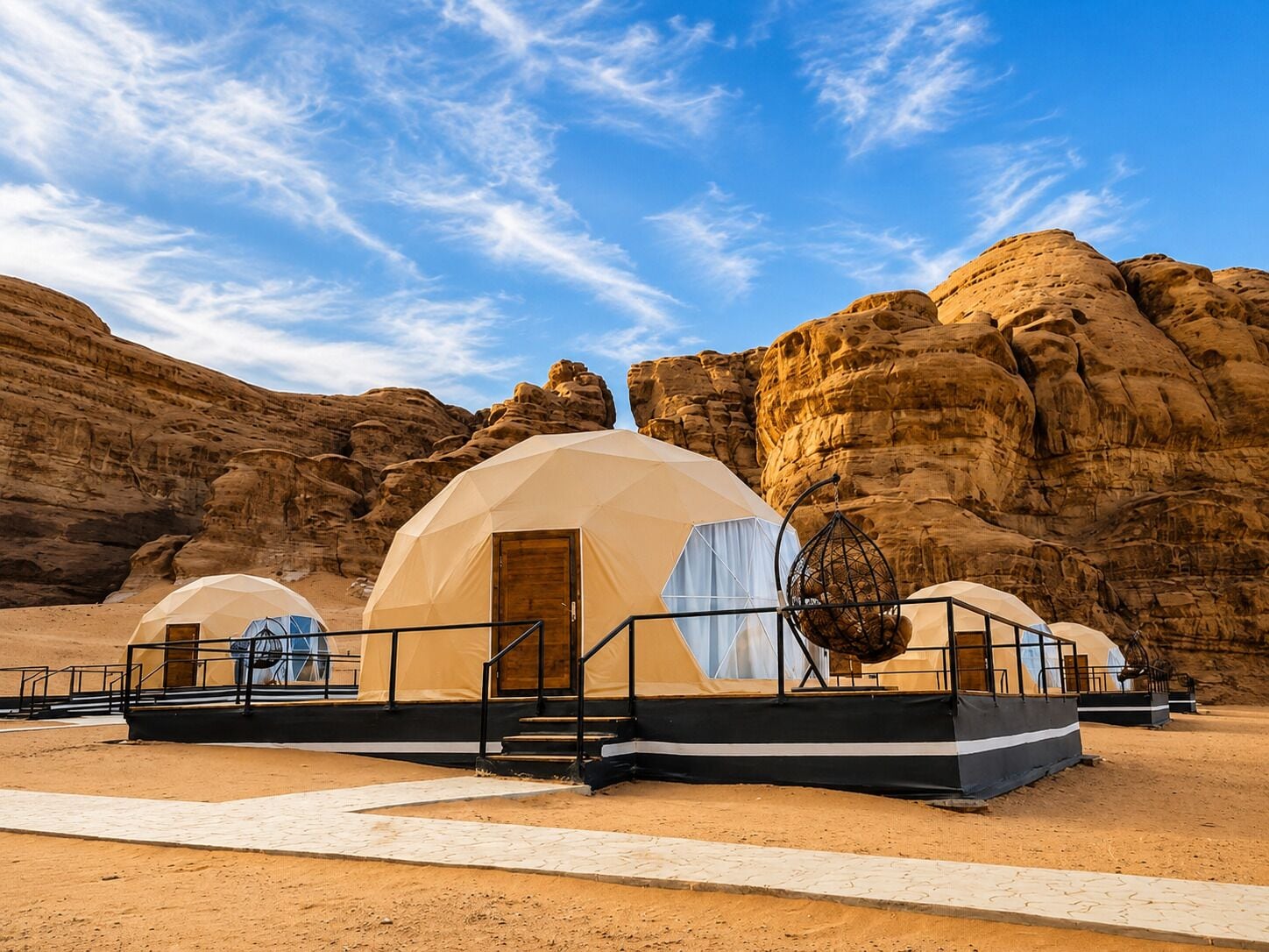 Paisaje desértico con tres tiendas en forma de cúpula, que tienen puertas y ventanas de madera, y están conectadas por una pasarela.