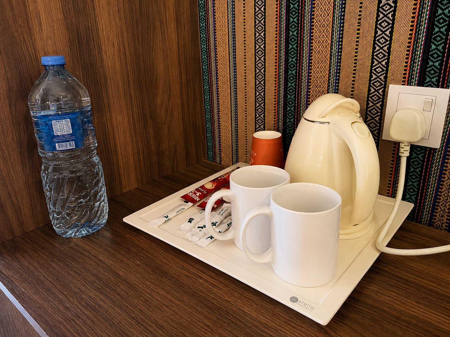 Una mesa de madera con una botella de agua, dos tazas blancas, una cafetera y un enchufe.