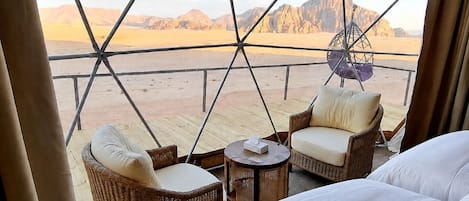 Una habitación con cama, dos sillas de mimbre, una mesita y vista a un paisaje desértico a través de una estructura de ventana geométrica.