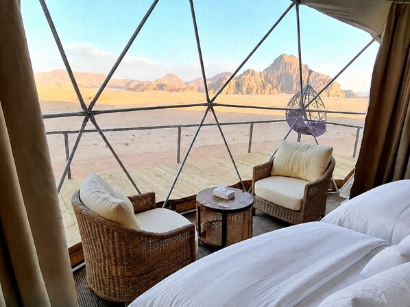 A room with a bed, two wicker chairs, a small table, and a view of a desert landscape through a geometric window structure.