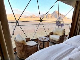 Una habitación con cama, dos sillas de mimbre, una mesita y vista a un paisaje desértico a través de una estructura de ventana geométrica.