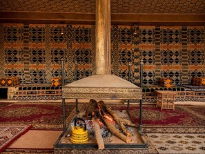 A traditional room with patterned walls, a central pillar, a fire pit with logs, and a yellow kettle. - Hasan Zawaideh Luxury Camp 2 (Wadi Rum)