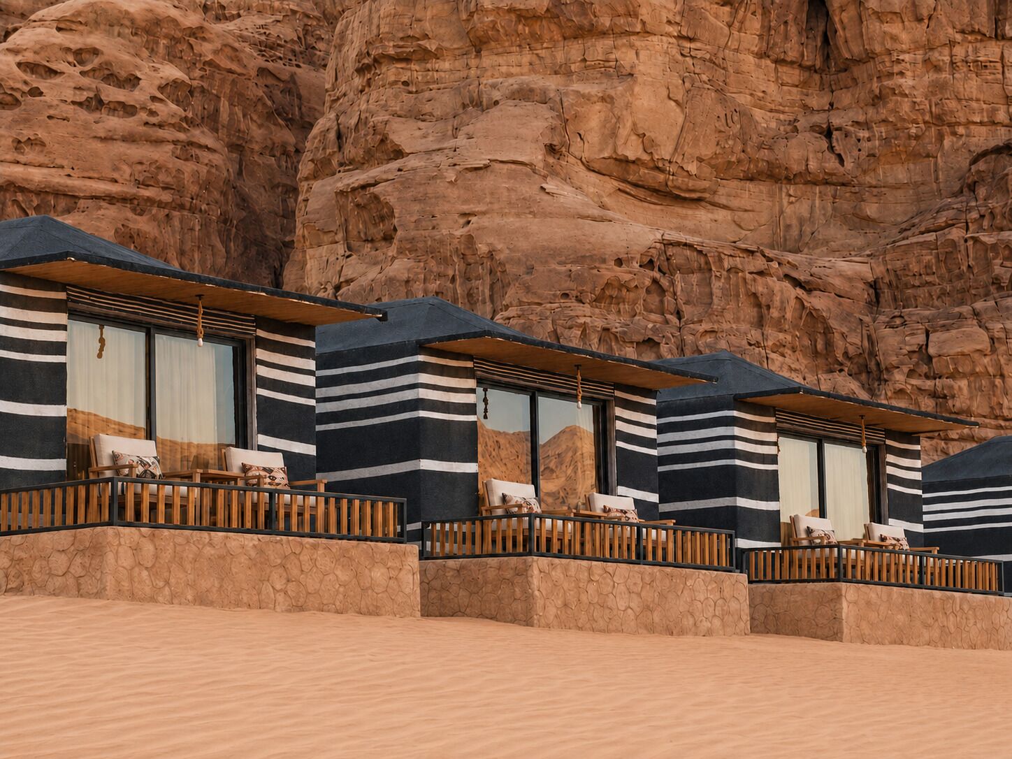 A row of modern, striped-roofed cabins with outdoor seating, set against a rocky cliff backdrop.