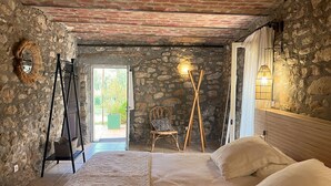 A rustic room with a stone wall, a bed with white linens, a wicker chair, and a wooden floor. -  Bastide 1830, Domaine de la Bastidonne 4* Superb view of le Luberon Heated Pool (Céreste)