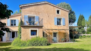 A two-story house with a balcony, a small window with a vase, and a covered patio area with chairs. -  Bastide 1830, Domaine de la Bastidonne 4* Superb view of le Luberon Heated Pool (Céreste)