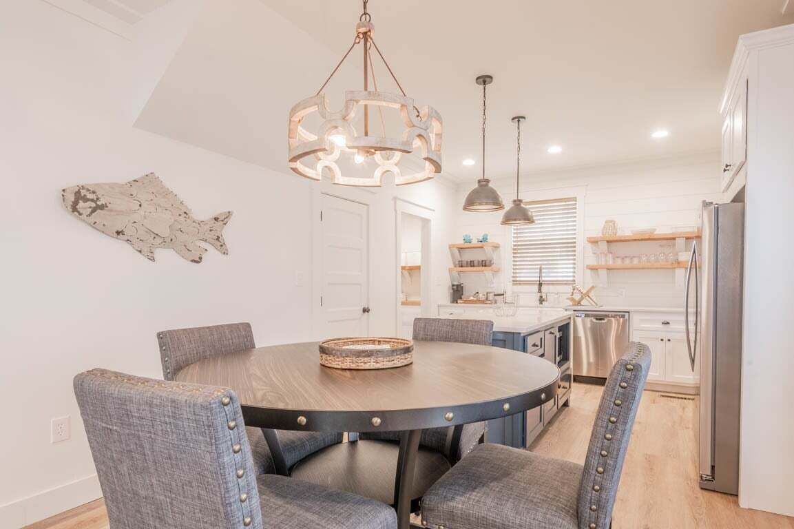 A dining area with a round table, grey upholstered chairs, a chandelier, and a kitchen in the background.