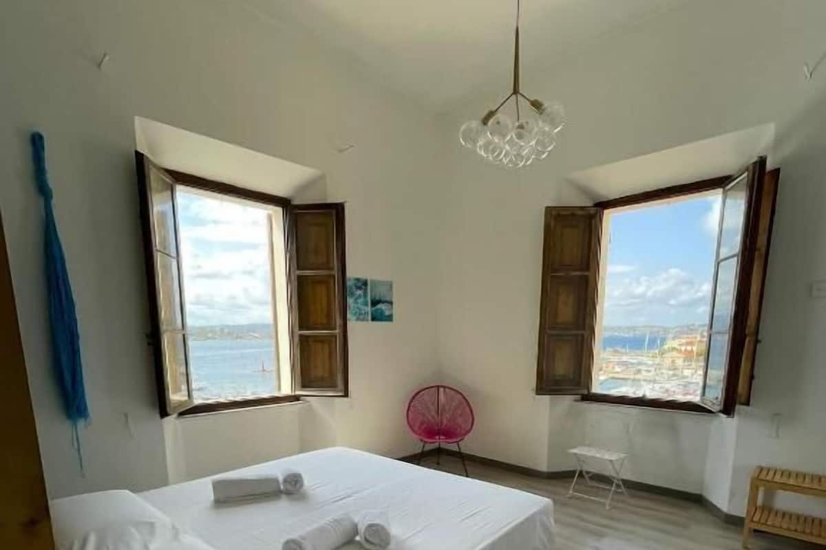 A bedroom with a bed, two windows, a chair, and a hanging light fixture.