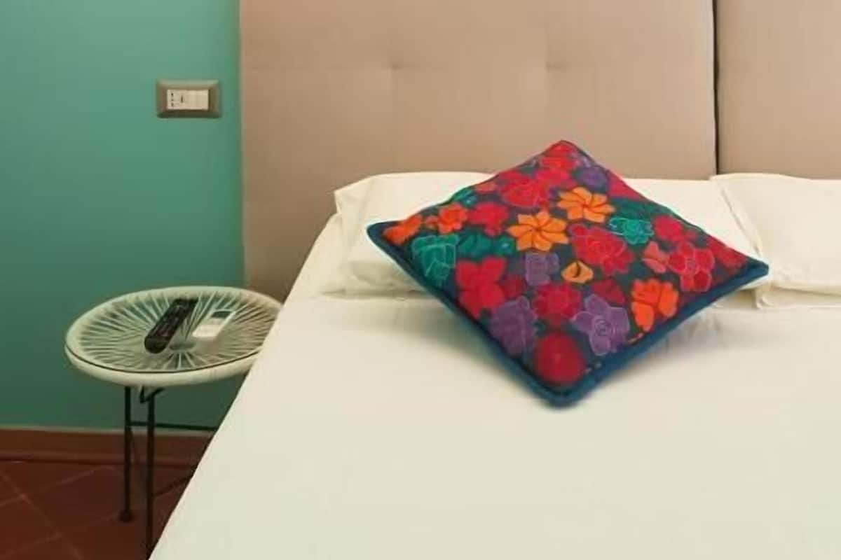 A bedroom with a teal wall, a white bed, a colorful pillow, a hanging light bulb, and a small white chair.
