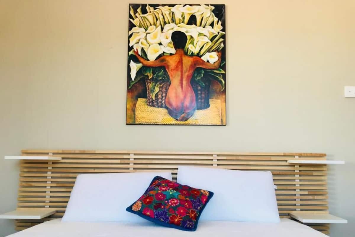 A hotel room with a bed, a colorful pillow, and a painting of a woman with flowers on the wall.