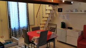 Una habitación acogedora con una mesa de comedor, sillas, un sillón rojo, un radiador, una estantería con utensilios de cocina y una ventana con cortinas.