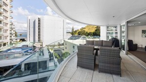 A modern balcony with a glass railing, a seating area with wicker furniture, and a view of a mountainous landscape. - A strand Of Paradise, Oceanside & Views with pool (Mount Maunganui)