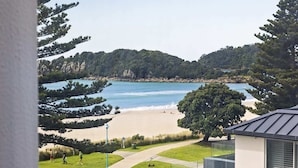 A beach view with a pathway leading to the shore, surrounded by greenery and a building with a sloped roof. - A strand Of Paradise, Oceanside & Views with pool (Mount Maunganui)