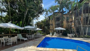 Una piscina con piso de baldosas, rodeada de mobiliario exterior con sombrillas y abundante follaje.