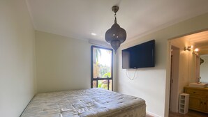 Un dormitorio con cama, televisor montado en la pared, ventana y baño con lavamanos y espejo.
