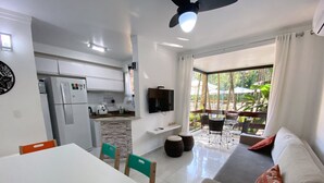 Una cocina moderna con refrigerador integrado, un comedor con mesa y sillas blancas, una sala de estar con sofá y televisor, y un balcón con mobiliario exterior.