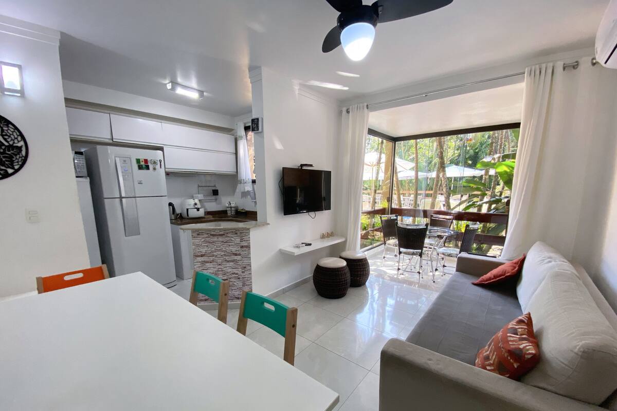 A modern kitchen with a built-in fridge, a dining area with a white table and chairs, a living area with a sofa and a TV, and a balcony with outdoor furniture.