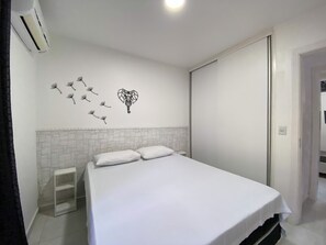 A bedroom with a bed, a white wall with a geometric design, a small white shelf, and a white tiled floor. - Riviera Green Residence (Bertioga)