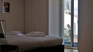 Une chambre à coucher avec un lit, une chaise et une vue sur un immeuble, visible à travers une porte vitrée.