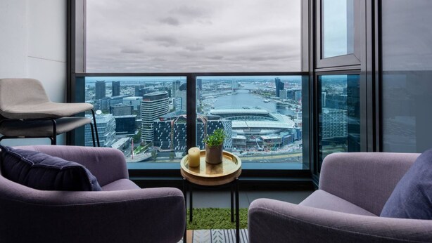A modern room with a large window offering a city view, two purple armchairs, a small round table with a plant and candle, and a chair. - Homely 1BR Apt Near Southern Cross Station w/ Pool (Melbourne)