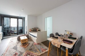 A modern, well-lit room with a large sliding glass door leading to a balcony, a small white bed, a dining table with chairs, and a colorful rug.