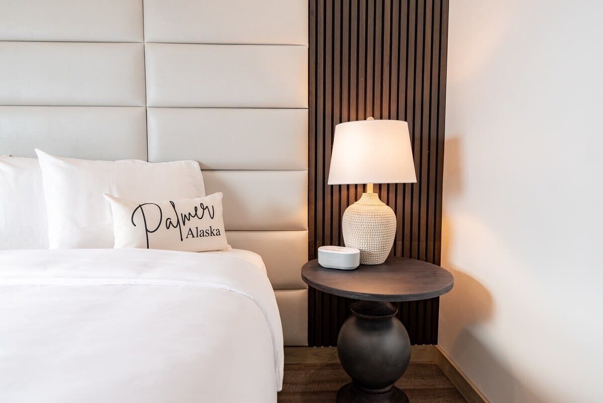 A hotel room with a bed, a pillow with "Palmer Alaska" text, a round table with a lamp, and a vertical wooden panel.