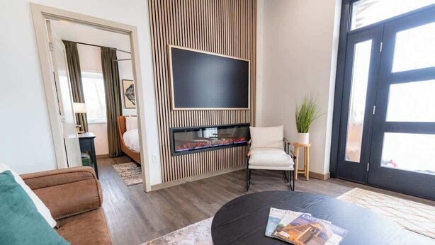 Un salon moderne avec un téléviseur à écran plat fixé sur un mur recouvert de panneaux en bois, un canapé marron, une table basse ronde avec un magazine et une porte à panneaux de verre.