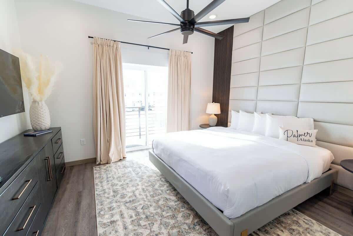 A modern bedroom with a large bed, a ceiling fan, a flat-screen TV, and a view of the outdoors.