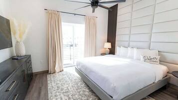 A modern bedroom with a large bed, a ceiling fan, a flat-screen TV, and a view of the outdoors.