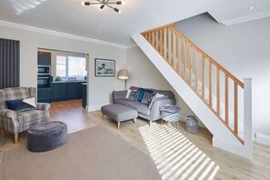 A modern living room with a staircase, a sofa set, and a kitchen visible through an open door.
