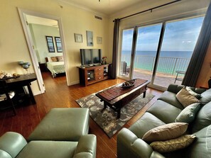 A living room with a sofa, a coffee table, and a TV. There is a balcony with a view of the ocean.