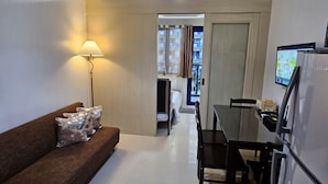 A compact living space with a sofa, a dining table, chairs, a refrigerator, and a lamp. - Sea Residences - Condo R Us (Pasay)