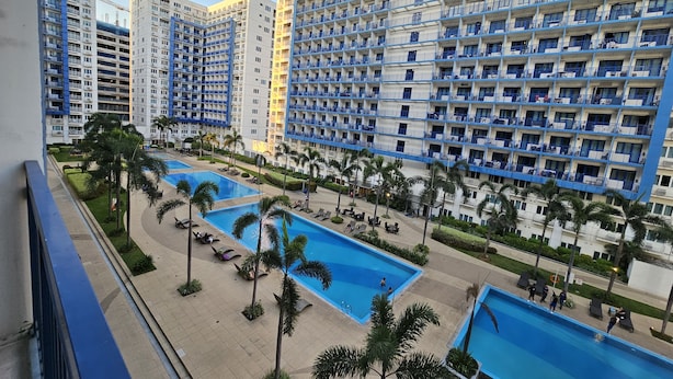 A high-rise hotel with multiple balconies, a swimming pool area with lounge chairs, and palm trees. - Sea Residences - Condo R Us (Pasay)