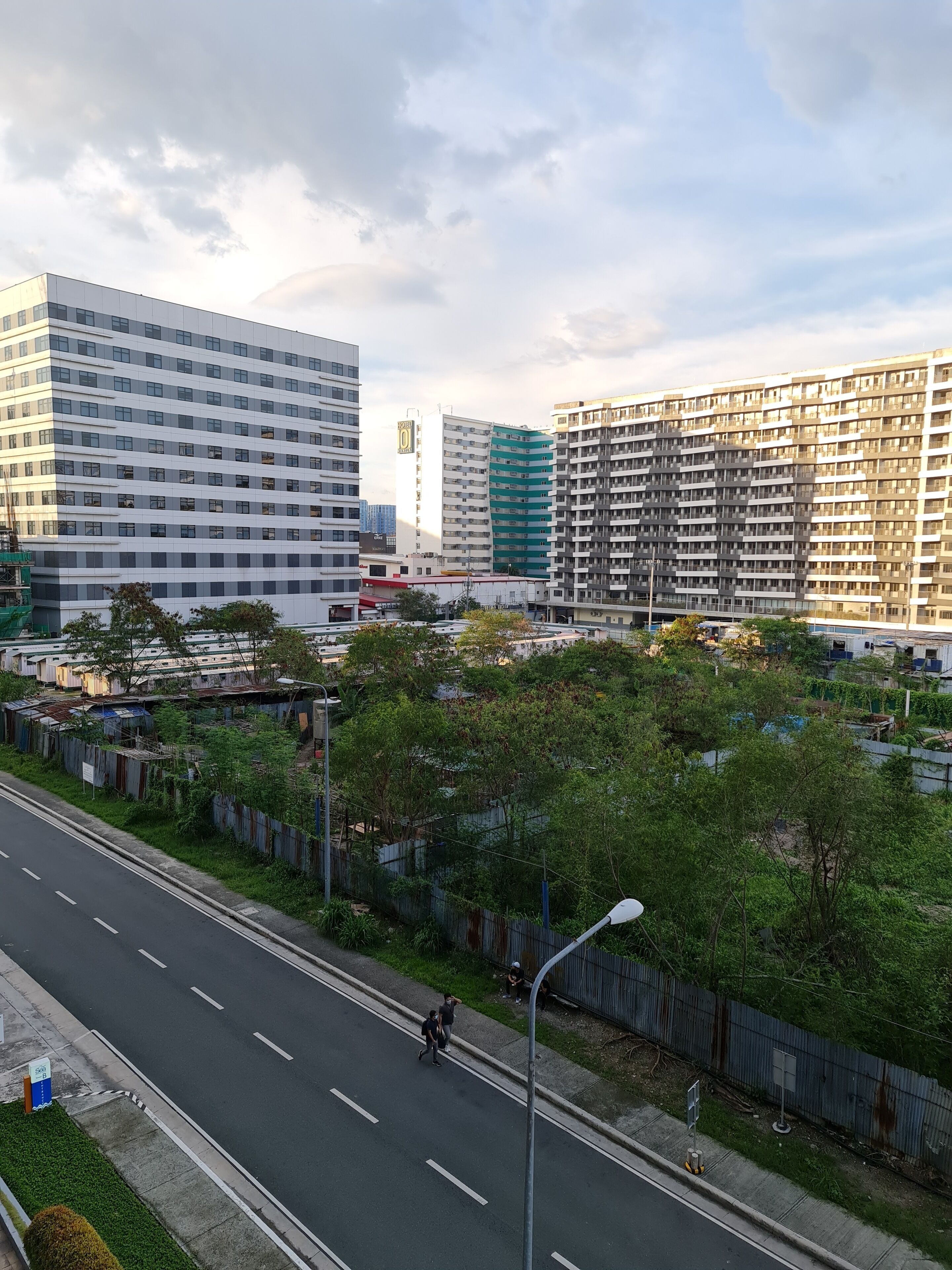 A modern urban street with high-rise buildings, a clear road, and greenery along the sidewalk.