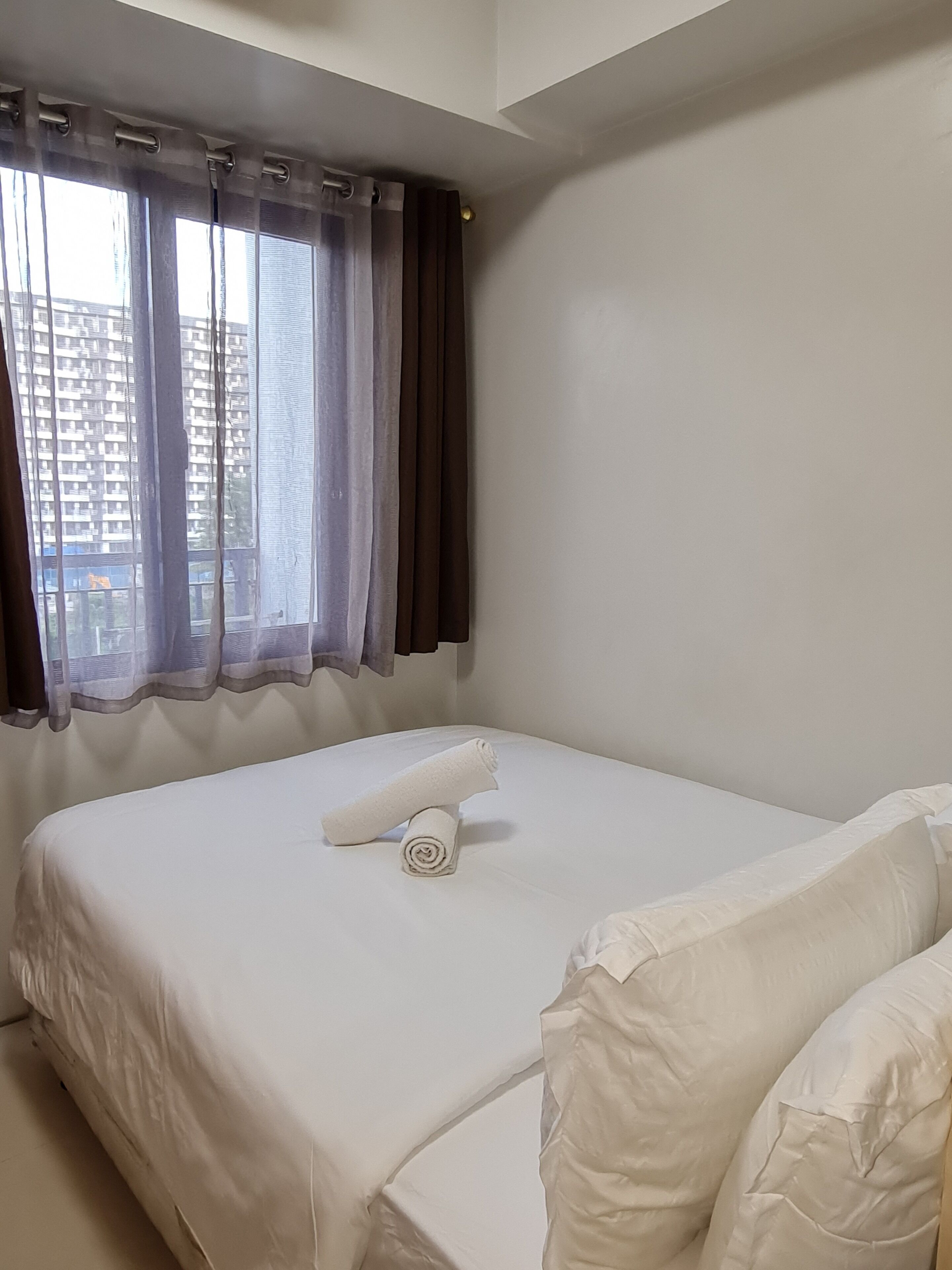 A neatly made bed with white linens and a rolled towel, a window with curtains, and a view of a building outside.