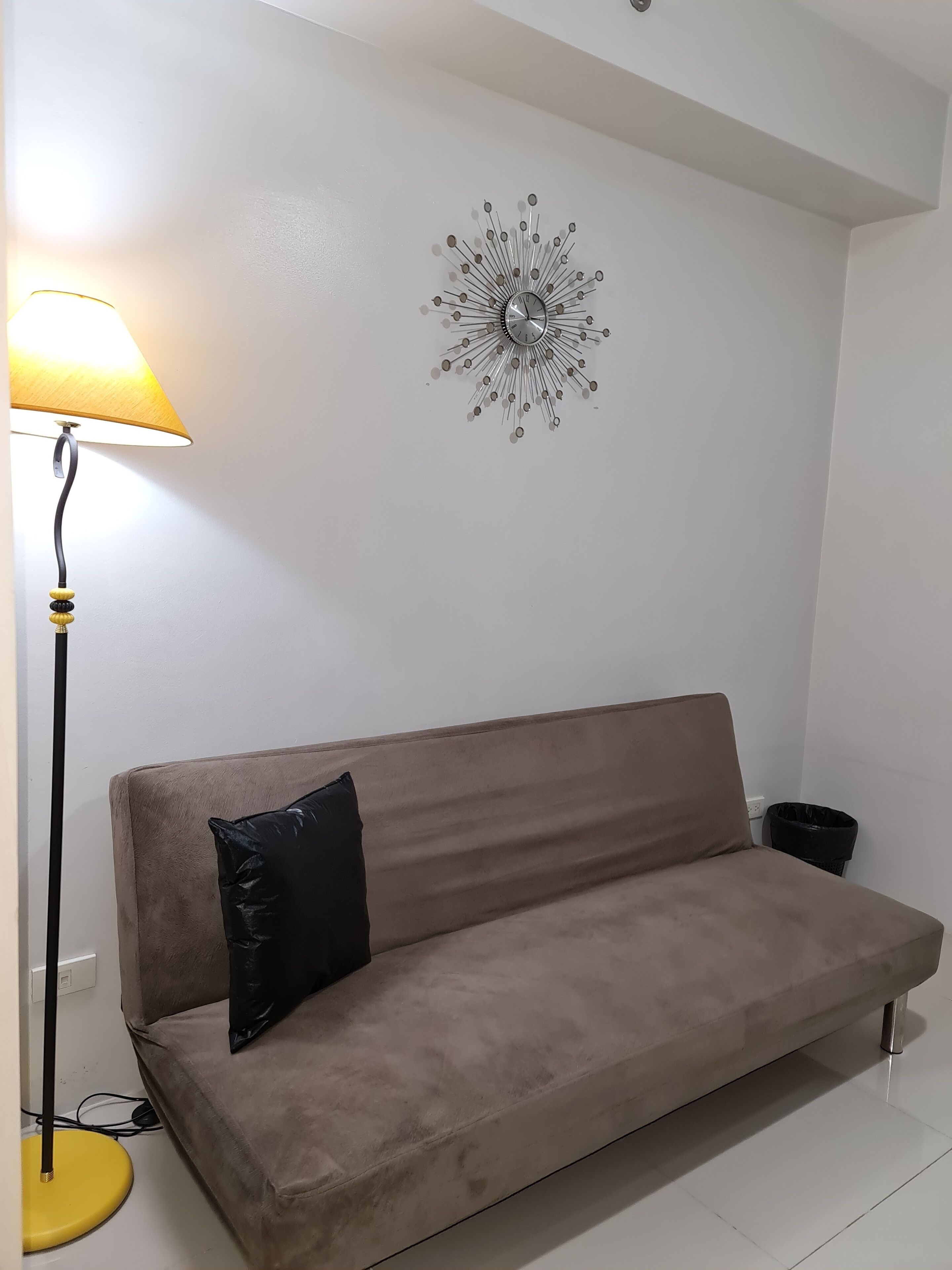 A beige sofa with a black pillow, a floor lamp with a yellow base, and a wall clock.