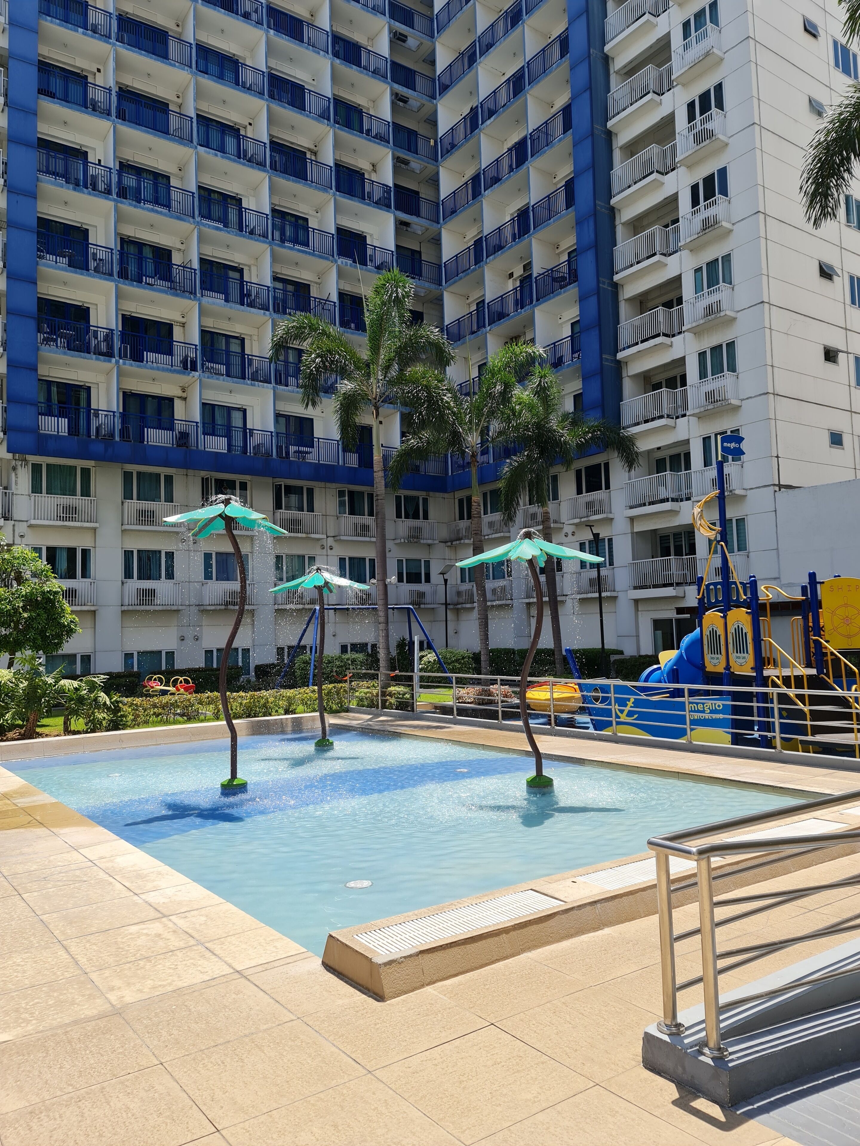 A swimming pool with three umbrellas, surrounded by palm trees and a multi-story building with balconies.