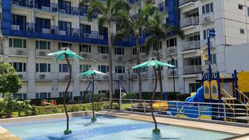 A swimming pool with three umbrellas, surrounded by palm trees and a multi-story building with balconies.