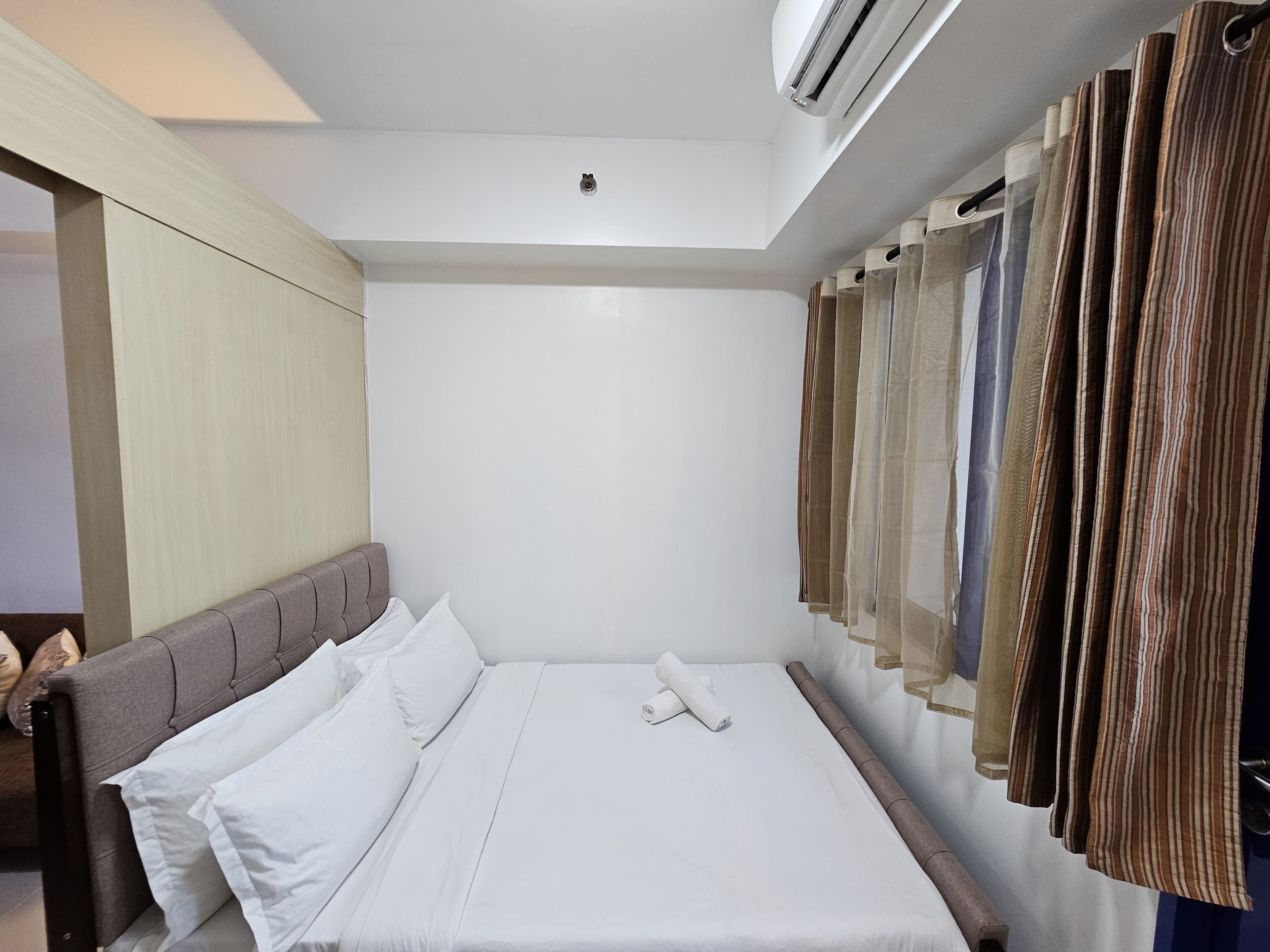 Une chambre d’hôtel comprenant un lit, une tête de lit, un climatiseur fixé au mur et une tringle à rideaux avec plusieurs rideaux.