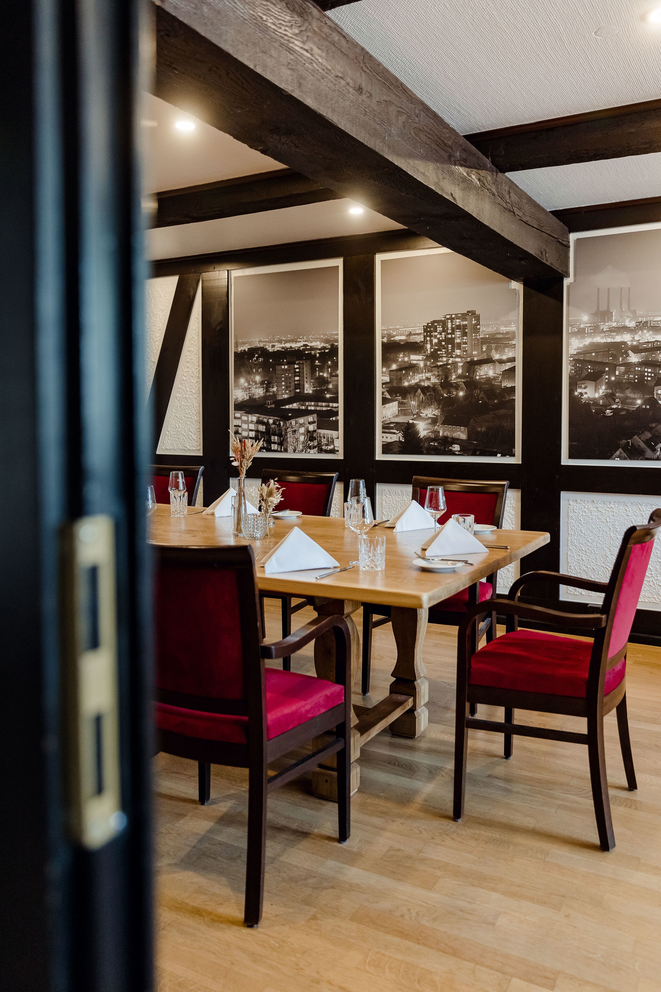 Un comedor con una mesa de madera dispuesta para cuatro personas, sillas tapizadas en rojo y fotografías en blanco y negro de un paisaje urbano en las paredes.