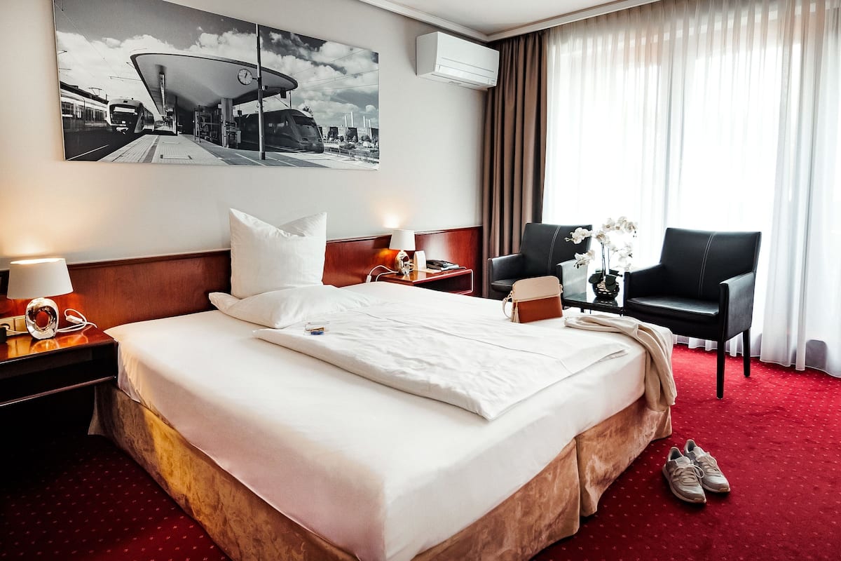 A hotel room with a large bed, a desk with a chair, a wall-mounted air conditioner, and a window with curtains.