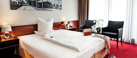 A hotel room with a large bed, a desk with a chair, a wall-mounted air conditioner, and a window with curtains.