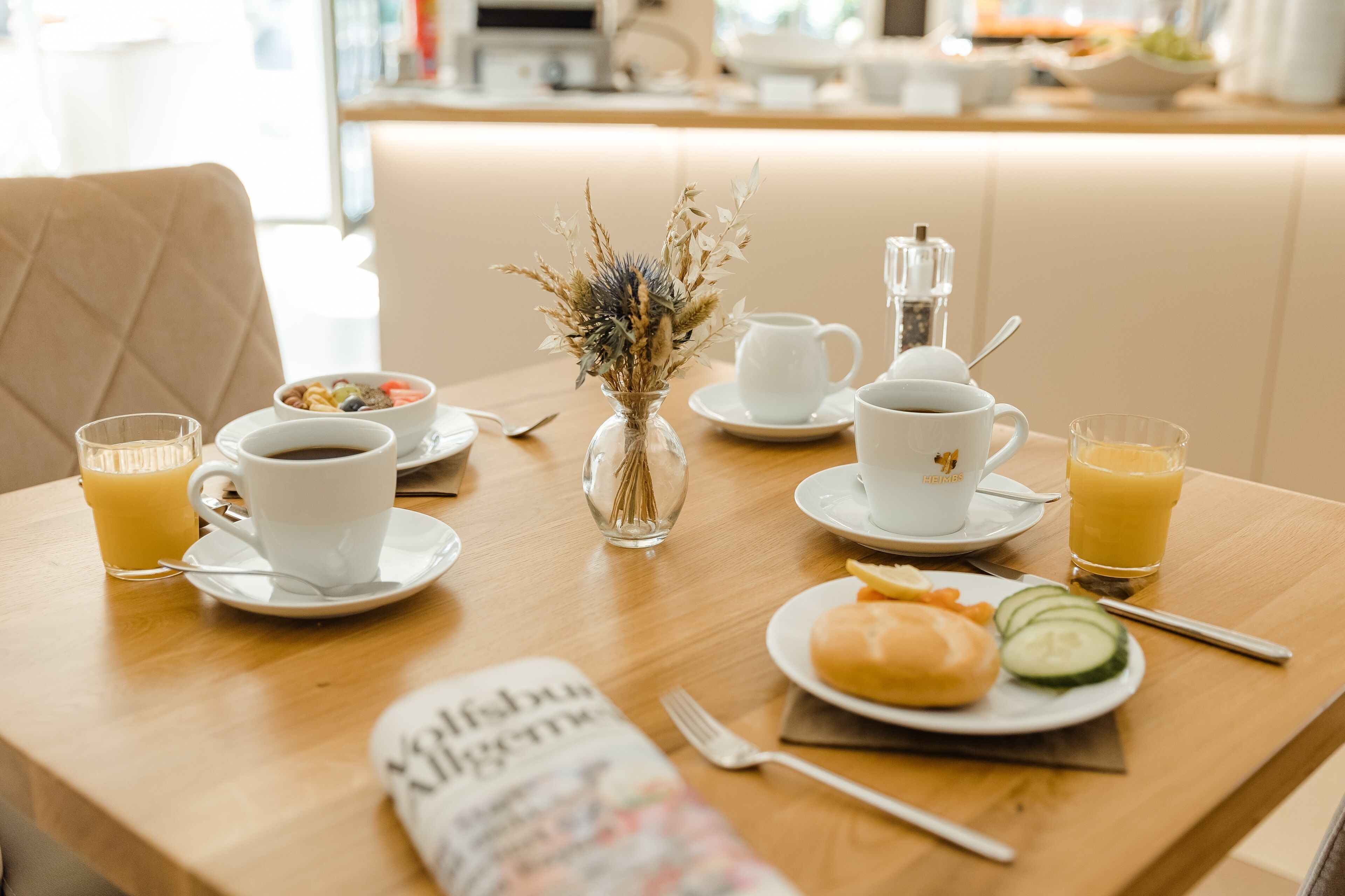 A breakfast table with a newspaper, coffee cups, a fruit bowl, and a muffin with a slice of cucumber.