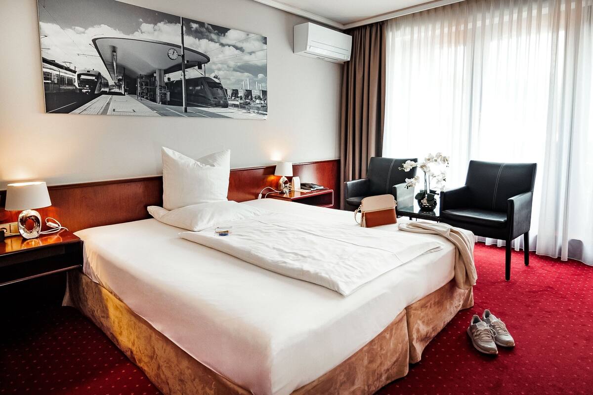 A hotel room with a large bed, a desk with a chair, a wall-mounted air conditioner, and a large window with curtains.