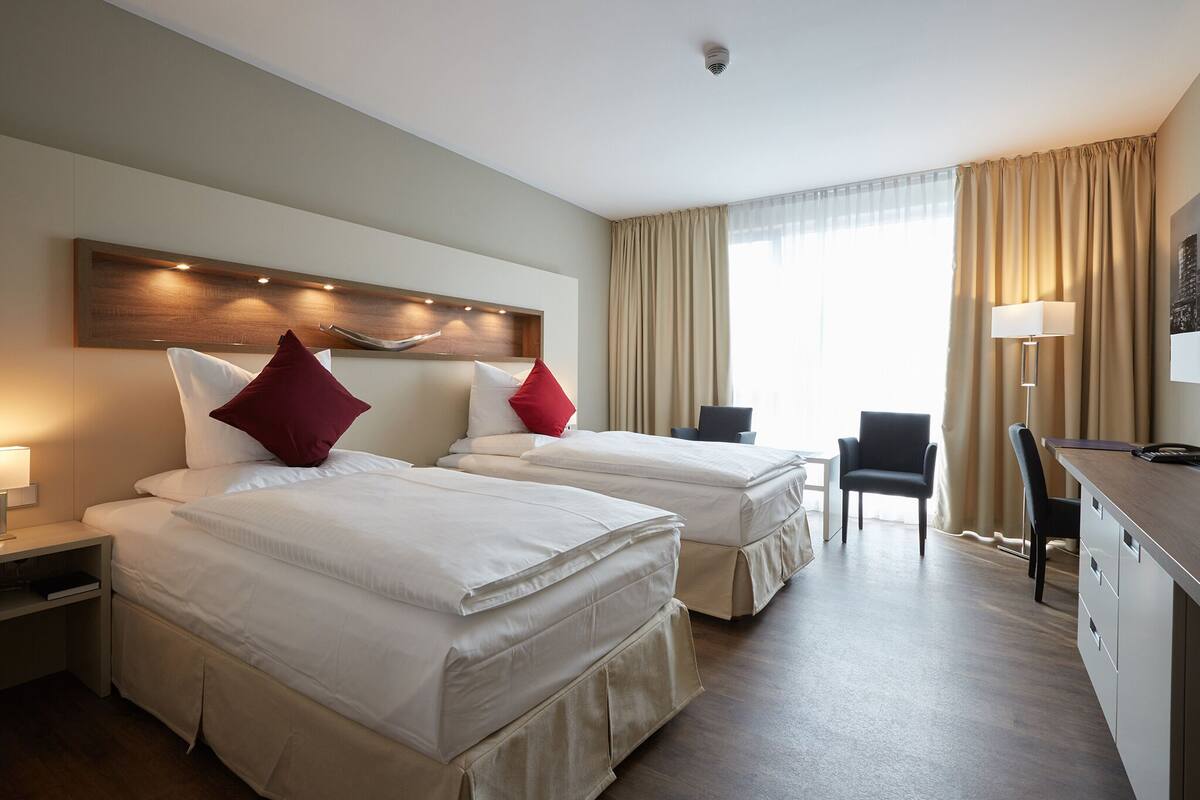 A hotel room with two beds, a desk, a chair, and a large window with curtains.