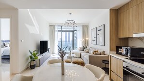 A modern hotel room with a dining area, kitchen, and a large window offering a city view.