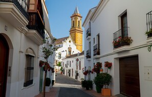 Eine enge Straße mit weißen Gebäuden, einer Kirche mit einem Glockenturm und Balkonen mit Blumen.