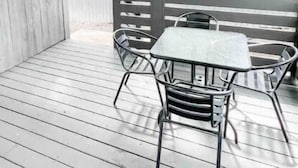 Une terrasse extérieure avec une plateforme en bois, une table et des chaises en métal, et un mur de briques en arrière-plan.